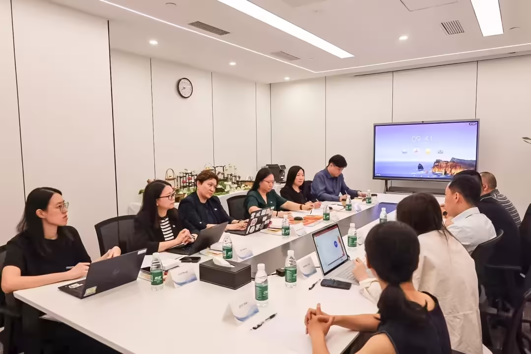 Participants from both sides seated around a white conference table in a modern meeting room, with laptops open and a large display screen visible at the far end.