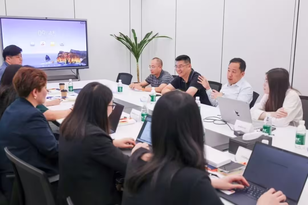 Several people engaged in animated discussion around a conference table, with one participant gesturing while speaking; a large screen and indoor plant are visible in the background.